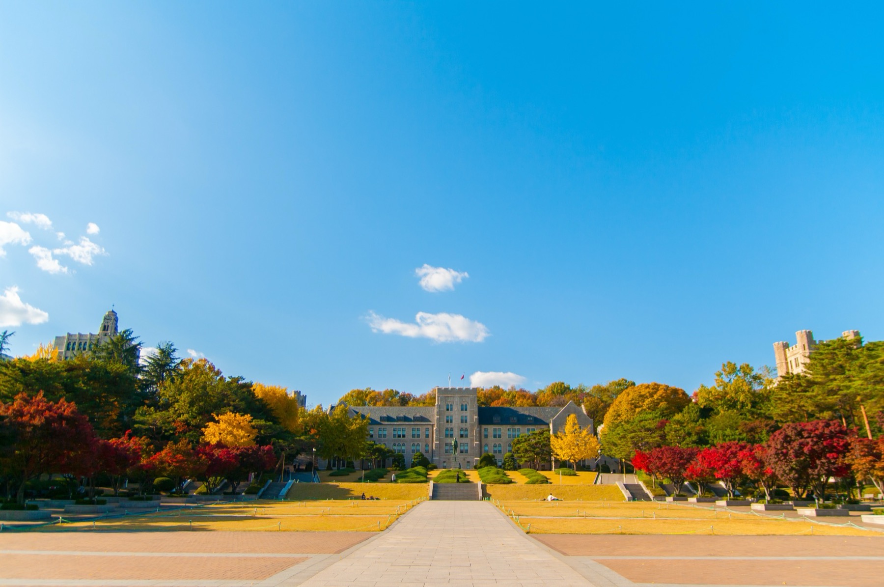 The stone main building of Korea University at the end of a long walkway, framed by red and yellow autumn trees under a blue sky.