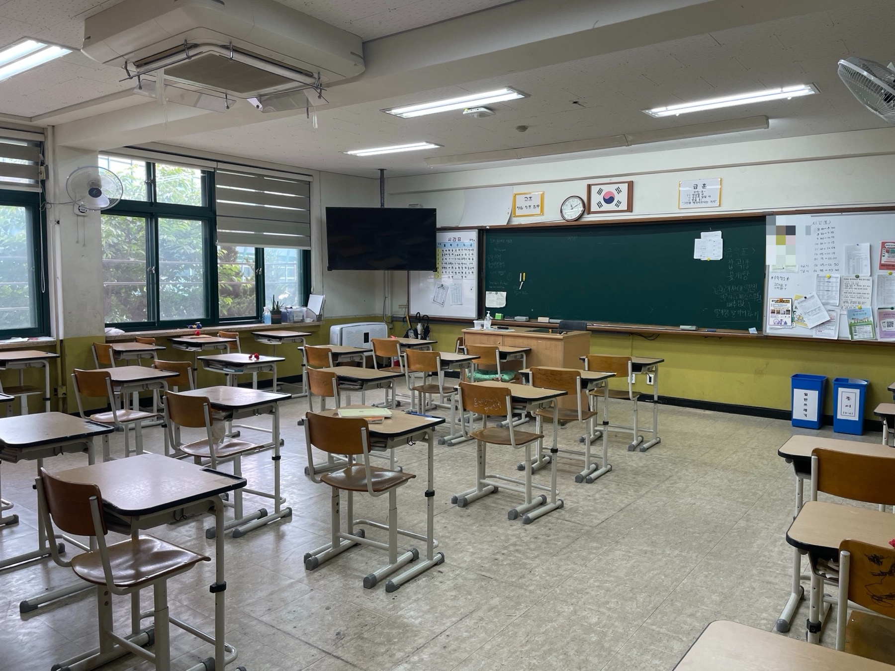 An empty South Korean classroom with rows of wooden desks and chairs, a green chalkboard, a whiteboard of notices and a Korean flag above the board.