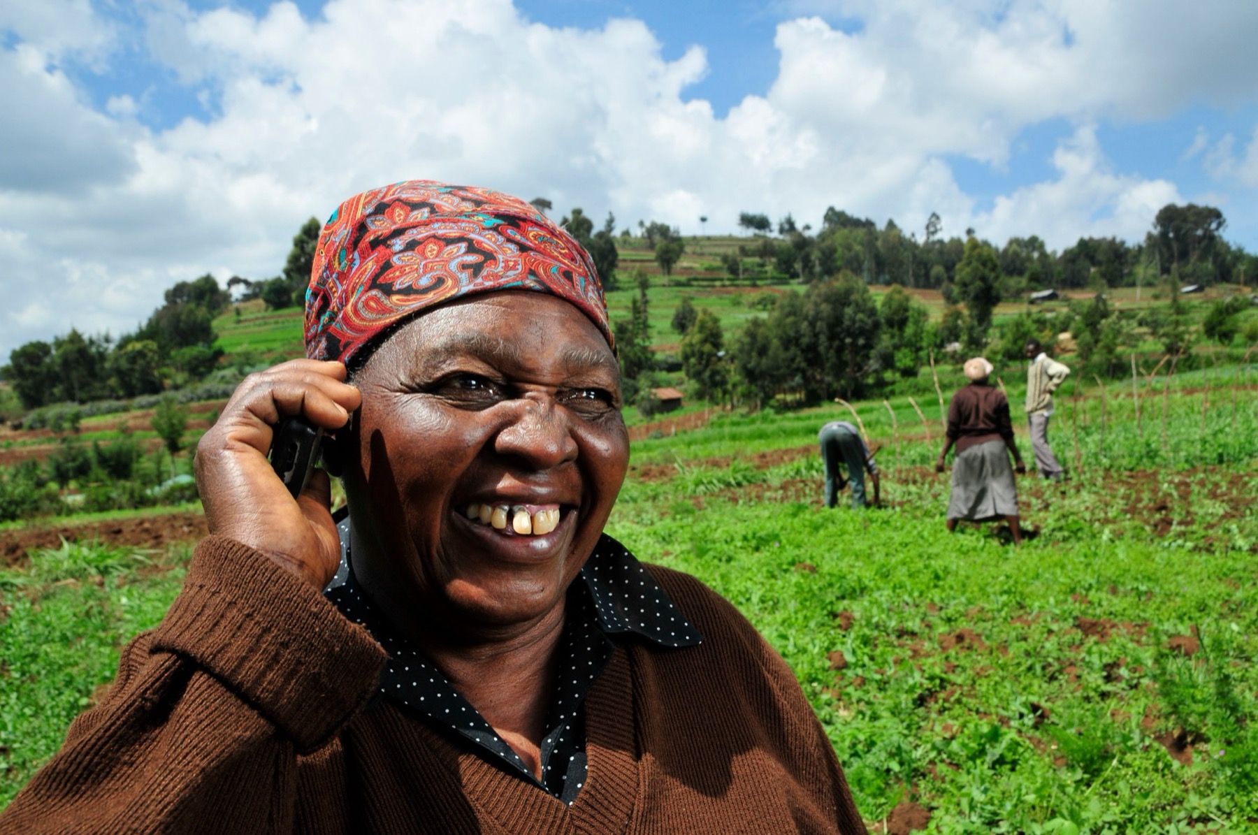 A smiling older woman in a red headscarf holds a mobile phone to her ear in a green field, with three people working the soil behind her.