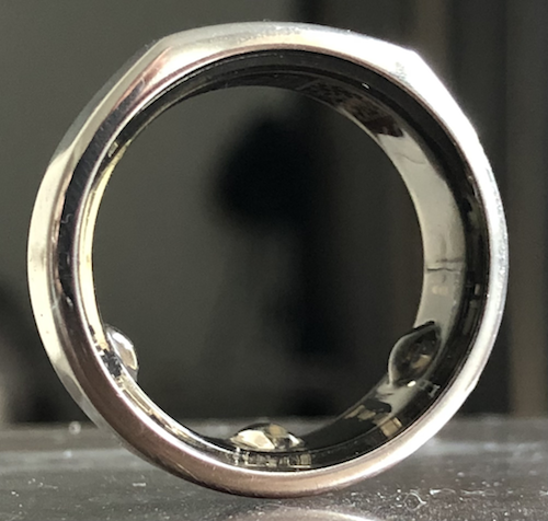A silver smart ring standing on its edge, with small sensor bumps visible on the inside of the band.