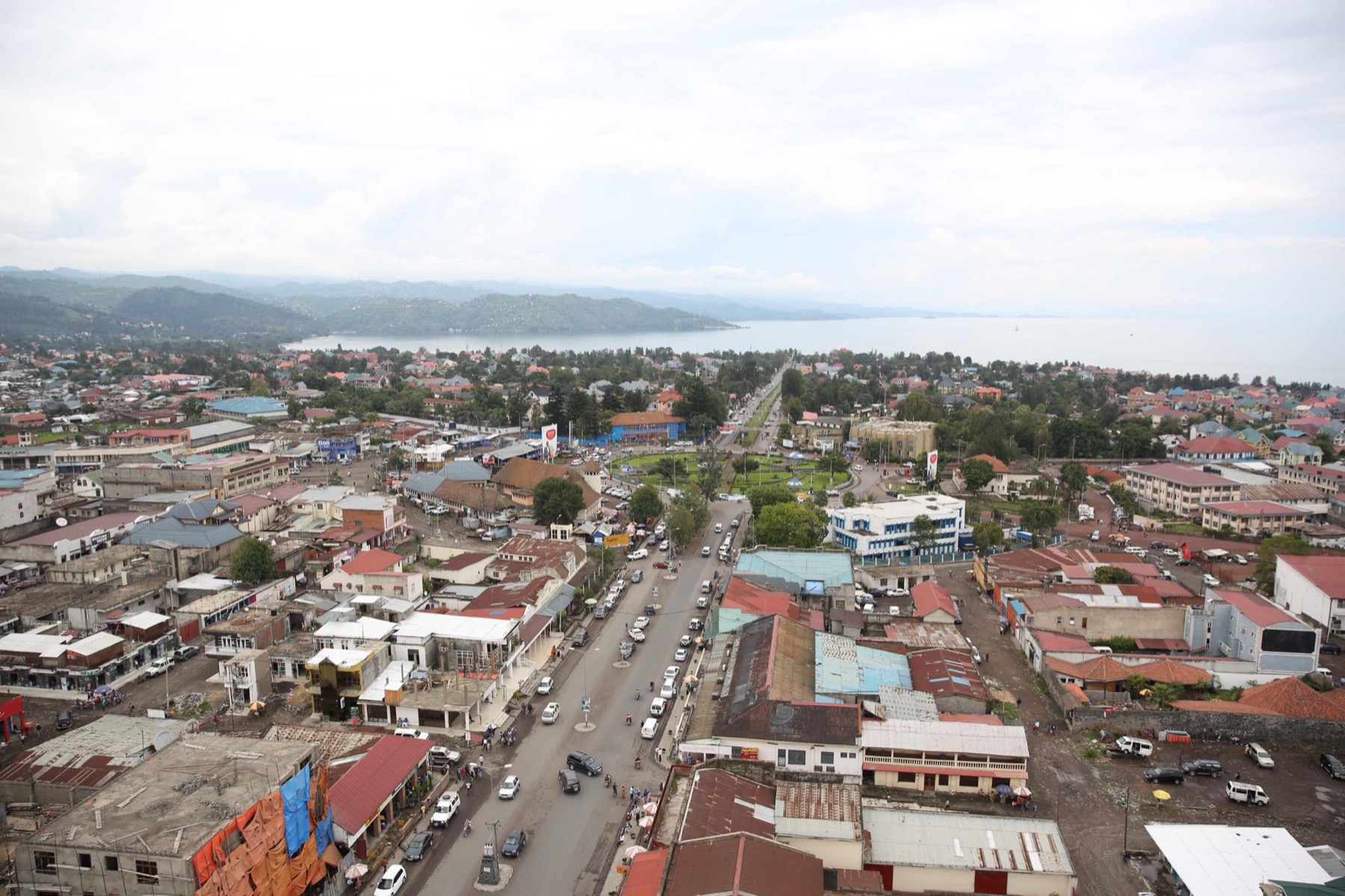 Aerial view of central Goma in 2015: a wide road with cars, a green roundabout, tin-roofed buildings and Lake Kivu with hills behind.