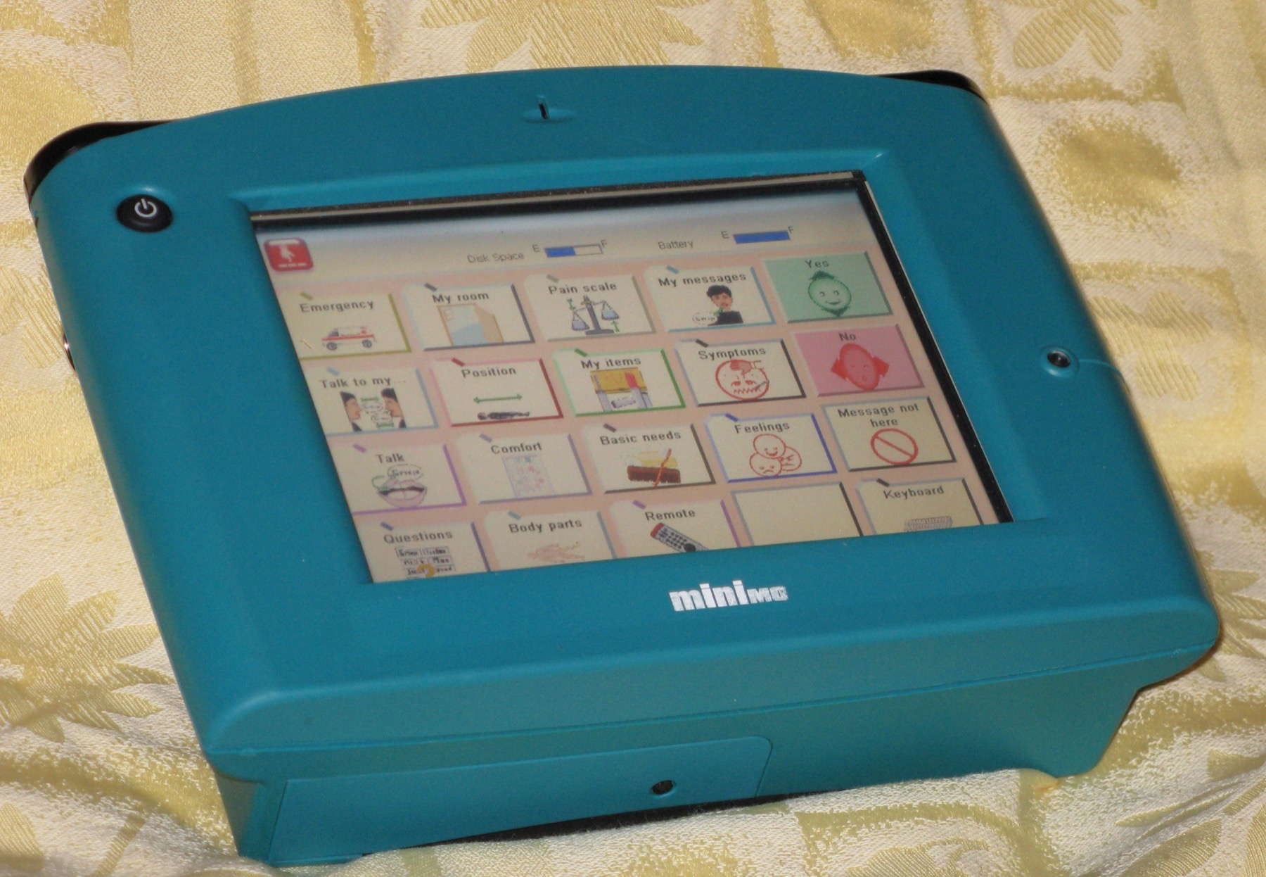 A teal speech generating device with a screen of picture buttons labelled Emergency, Talk, Feelings, Yes, No and Keyboard.