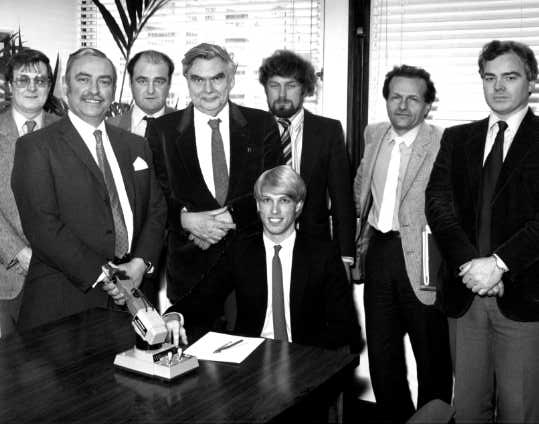 Black and white group photo of eight men in suits; a young fair-haired man sits at a table with a small robot arm model while seven men stand around him