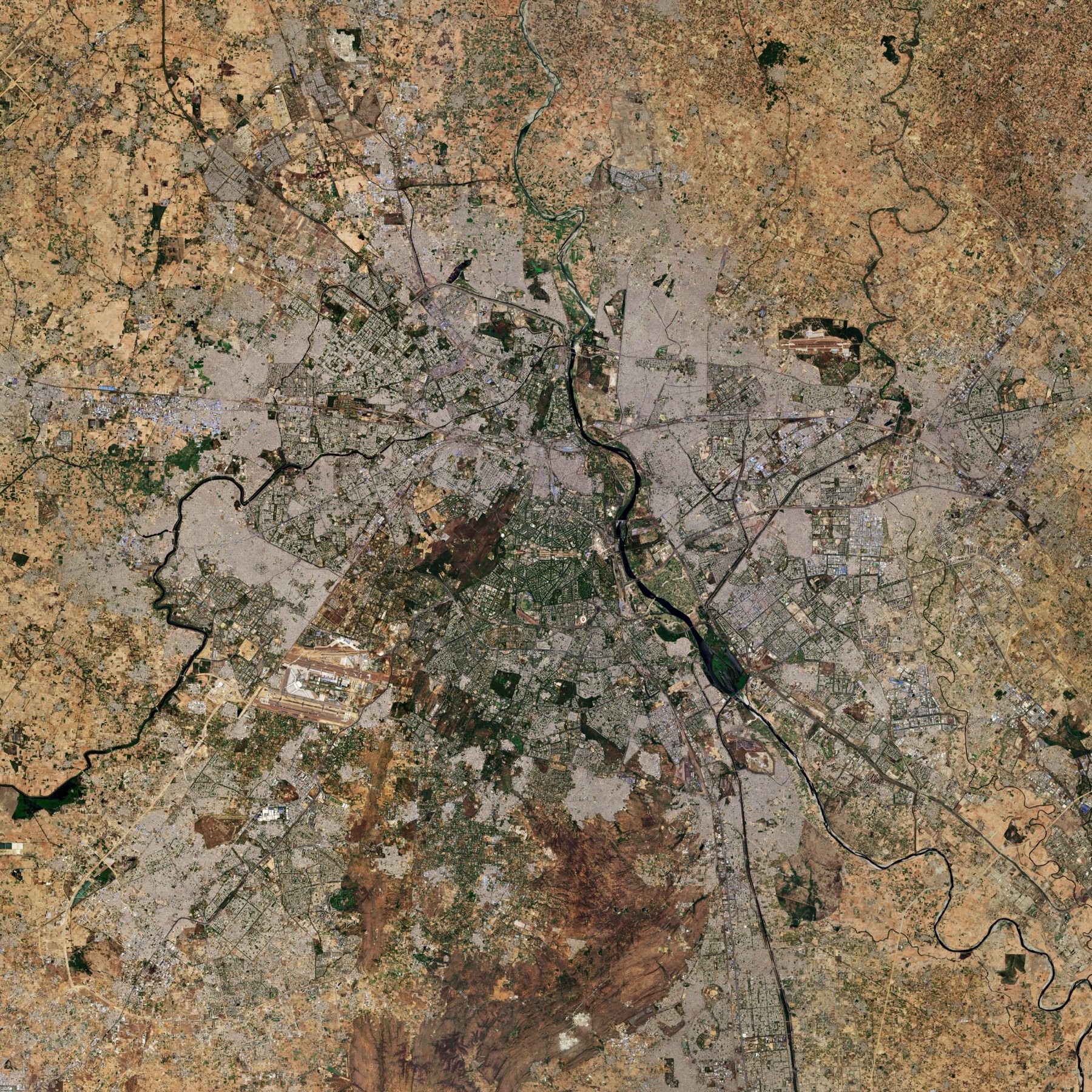 A true-color satellite view of the Delhi area: grey city districts, the dark line of the Yamuna River, airport runways and tan farmland at the edges.